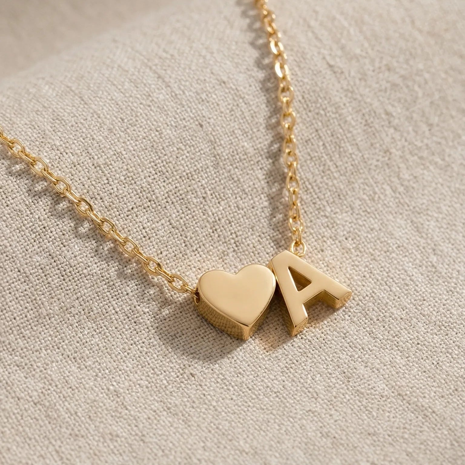 Gold necklace with a heart and letter 'A' pendant on a beige fabric background