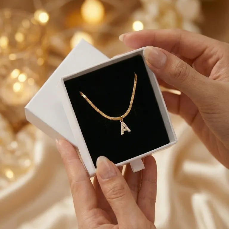 Gold necklace with a letter 'A' pendant in a white box held by hands against a warm, blurred background.